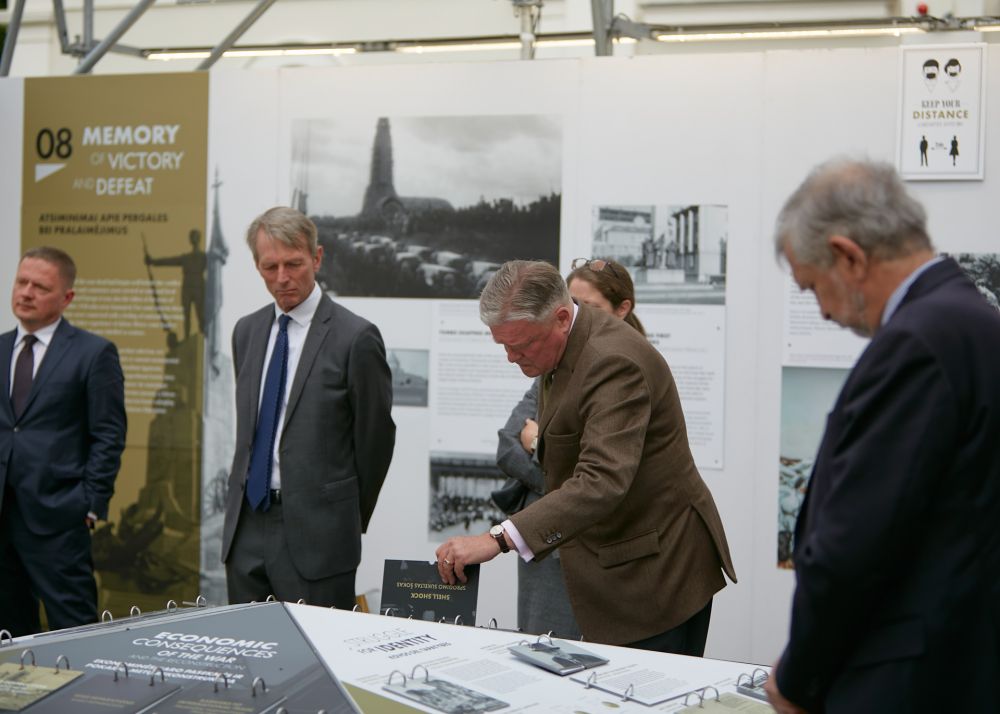 Interior of the installation of After the Great War exhibition. A man in the brown suit inspects closely the board with the infographics at the center, lifting one of the panels under which additional information is hidden. From the distance, four other people are watching attentively.