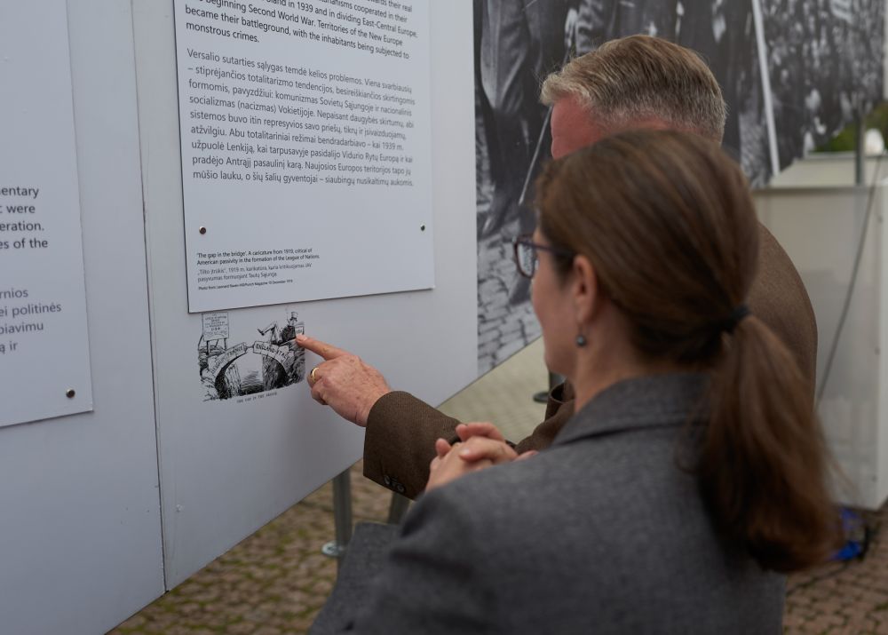 A man and a woman face the internal wall of the After the Great War exhibition. They are inspecting closely the photography presented there, towards which the man is pointing his finger.