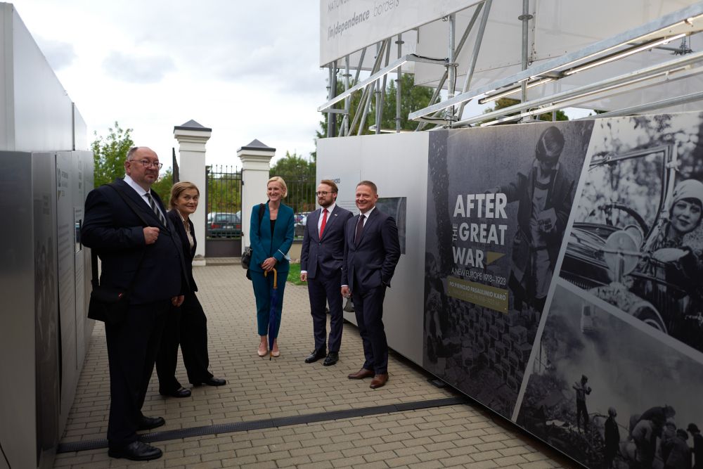 A group of five well-dressed people faces the camera smiling. They are standing in front of the entrance to the installation of the After the Great War exhibition, surrounded on both sides by its walls.
