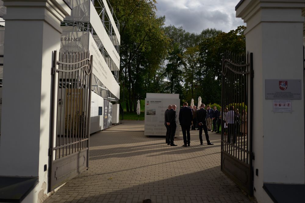 The open neo-classical gate reveals insides of the garden. In the centre of it stands the cube-like installation of After the Great War exhibition. It is surrounded by the groups of conversing guests.