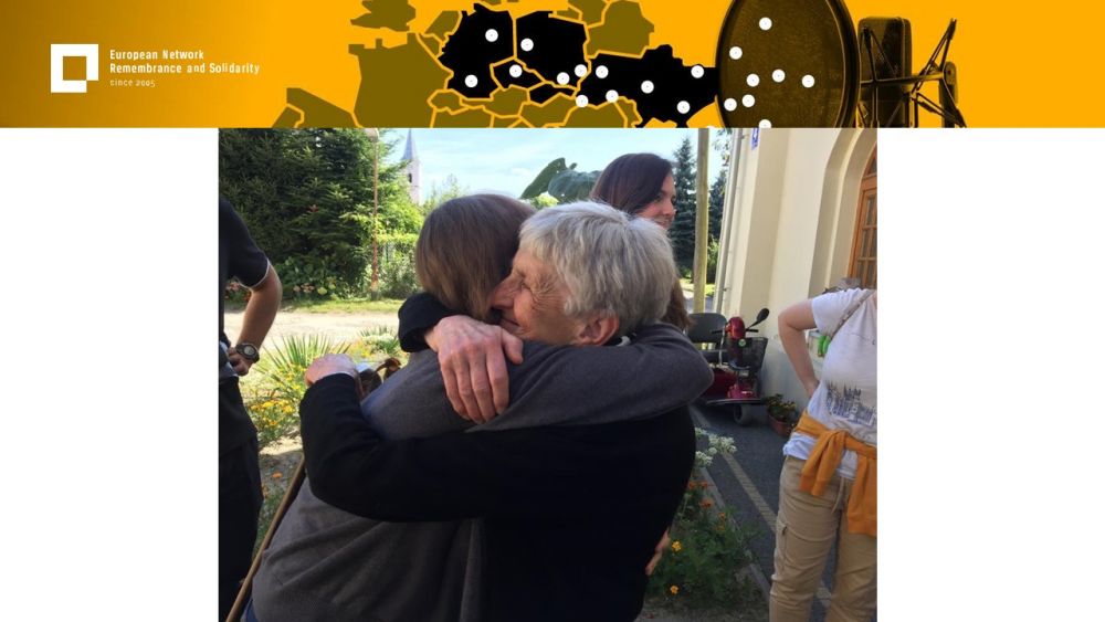 Presentation slide with a single photo in the center. In it, a young woman embraces old woman. They are standing outdoors. Greenery and other people in the background. Above all of it, a gold-yellow header with European Network remembrance and Solidarity.