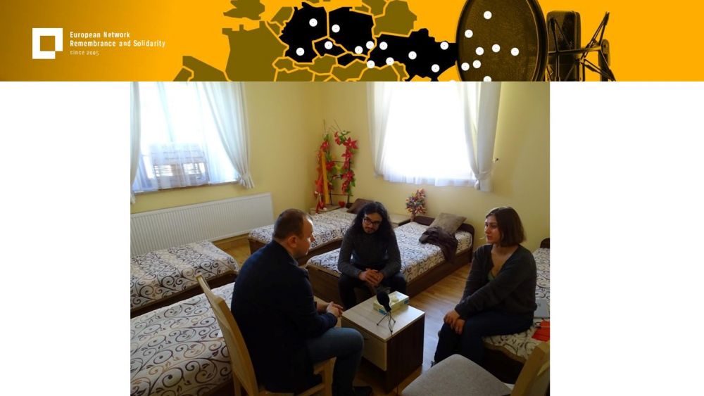 Presentation slide with a single photo in the center. In it, a hostel room with six beds in it. Two young people interview an adult man who sits in front of them. Above all of it, a gold-yellow header with European Network remembrance and Solidarity.