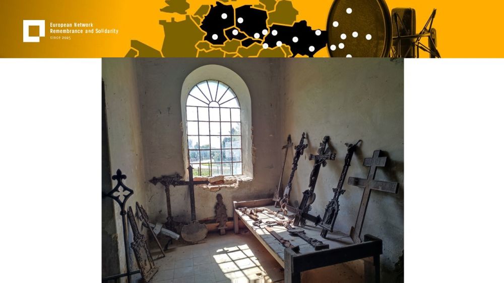 Presentation slide with a single photo in the center. In it, a bare room with a large window overlooking a village road. In the interior, multiple orthodox crosses out of iron lay without any visible order.