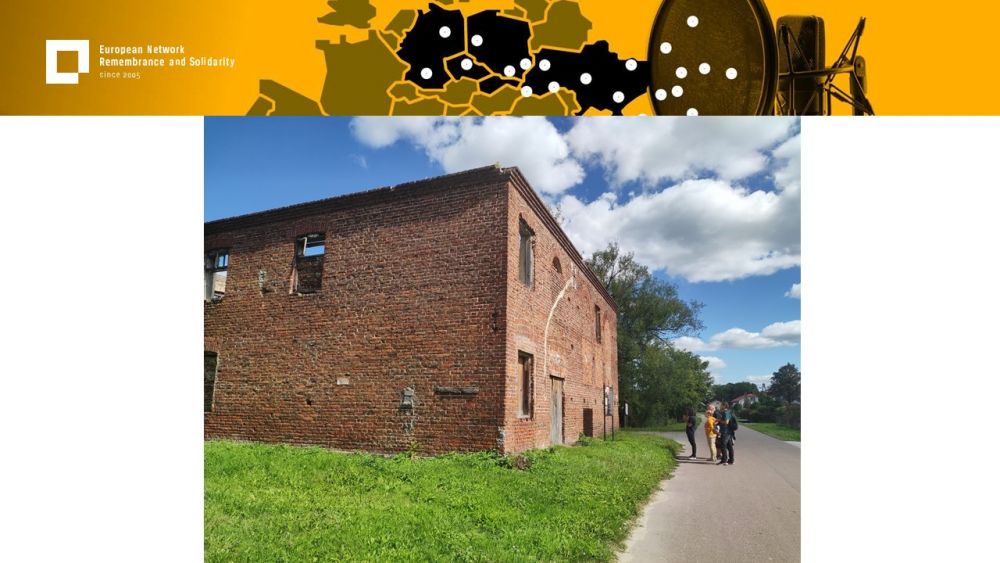 Presentation slide with a single photo in the center. The photo shows a group of onlookers stopping in front of a large abandoned building made of red brick. Above all of it, a gold-yellow header with European Network remembrance and Solidarity.