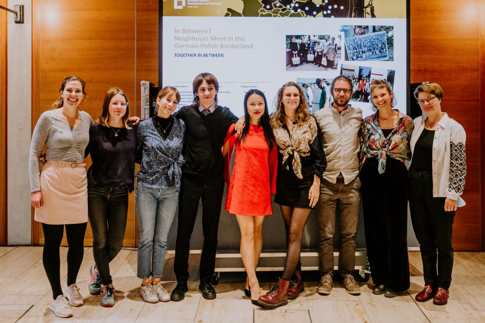 Interior, bright space with wooden panels. A group of nine people, of various age and gender stands in front of the screen with the presentation of their final In Between? project. Most of them are high-school-aged. They are all facing the camera, smiling.