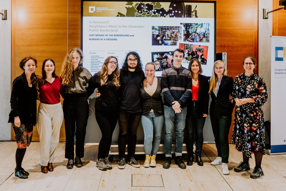 Interior, bright space with wooden panels. A group of ten people, of various ages and gender stands in front of the screen with the presentation of their final In Between? project. At least half of them are high-school-aged. They are all facing the camera, smiling.