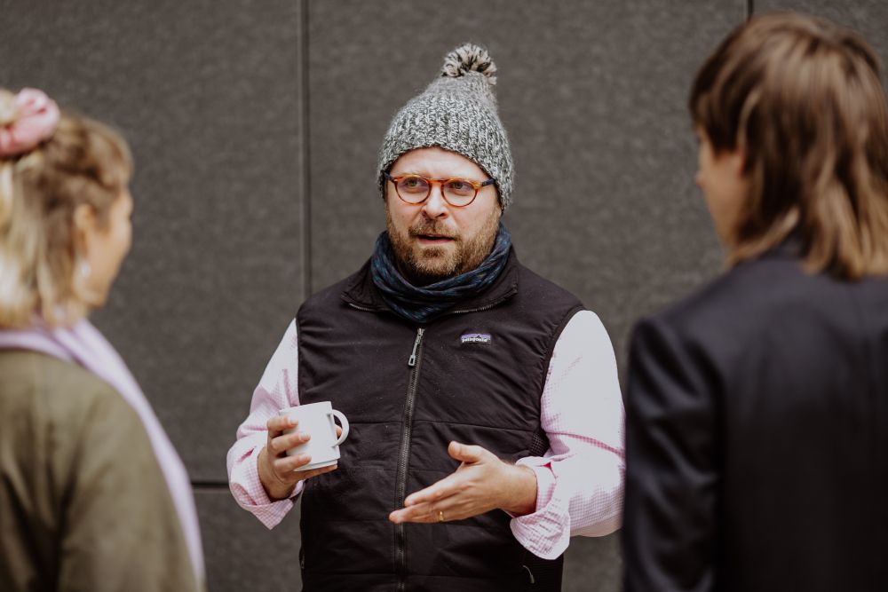 Interior. A man in a hat and a puffy vest holds a cup in his hand, facing the camera. He talks with two young people whose backs are out of focus.