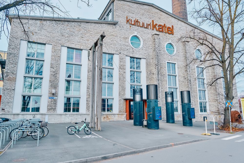 A large postindustrial building made of white brick with high windows. In front of it, bikestands, electric poles, and a leafless tree. On its facade, a large orange sign reads Kultuurikatel.