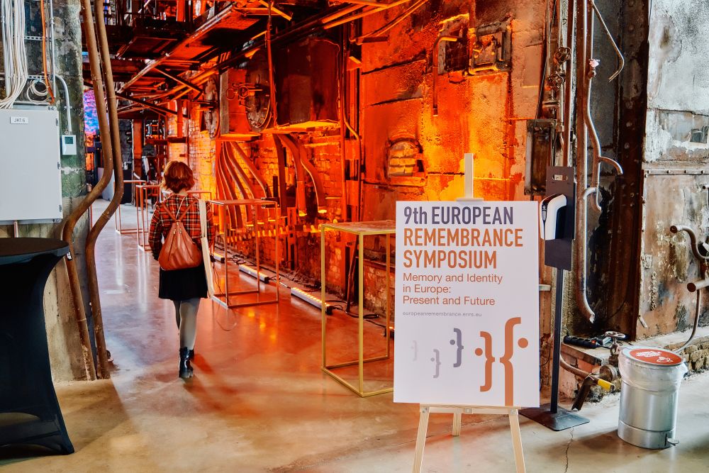 Interior. A spacious postindustrial hall is illuminated with orange lights. A woman, facing back the camera, walks through the hall. On her right stands the white board announcing the ninth European Remembrance Symposium in Tallinn.