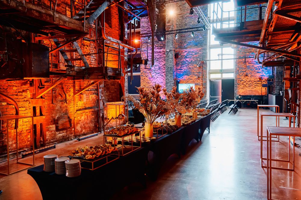 Interior. A spacious postindustrial hall is illuminated with orange lights. In a centre of the photo stands a buffet table filled with orderly prepared drinks and snacks. Further in the background, empty rows of auditorium chairs.