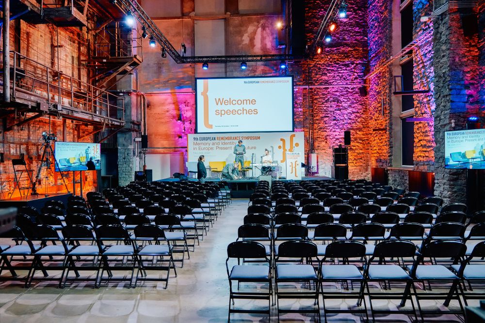 Interior. A spacious postindustrial hall is illuminated with orange lights. In the center of the photo, four people work to set up a stage for the European Remembrance Symposium. In front of them, many rows of foldable black chairs of the yet empty auditorium.