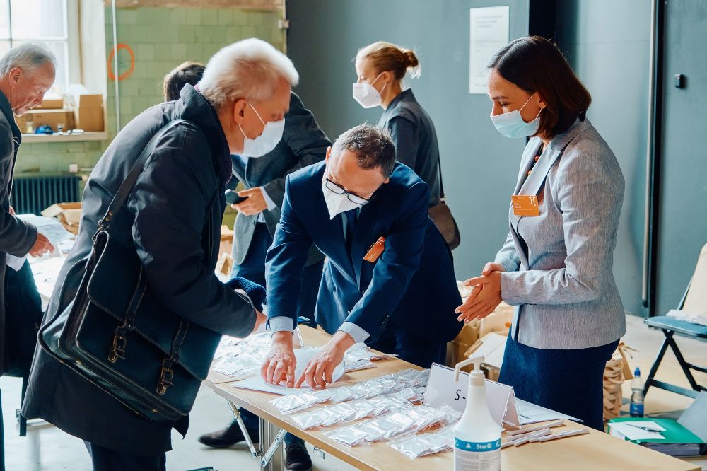 Interior. An older man leans over a table with rows of ID badges displayed in front of him. On the other side of the table, a well-dressed man and woman try to help him, looking for the appropriate badge.