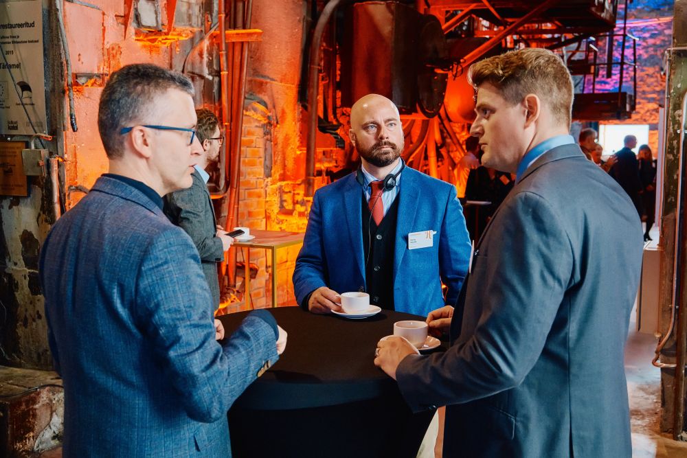 Interior. Three men, guests of the Symposium,  are engaged in a lively conversation. They stand by the high coffee table.