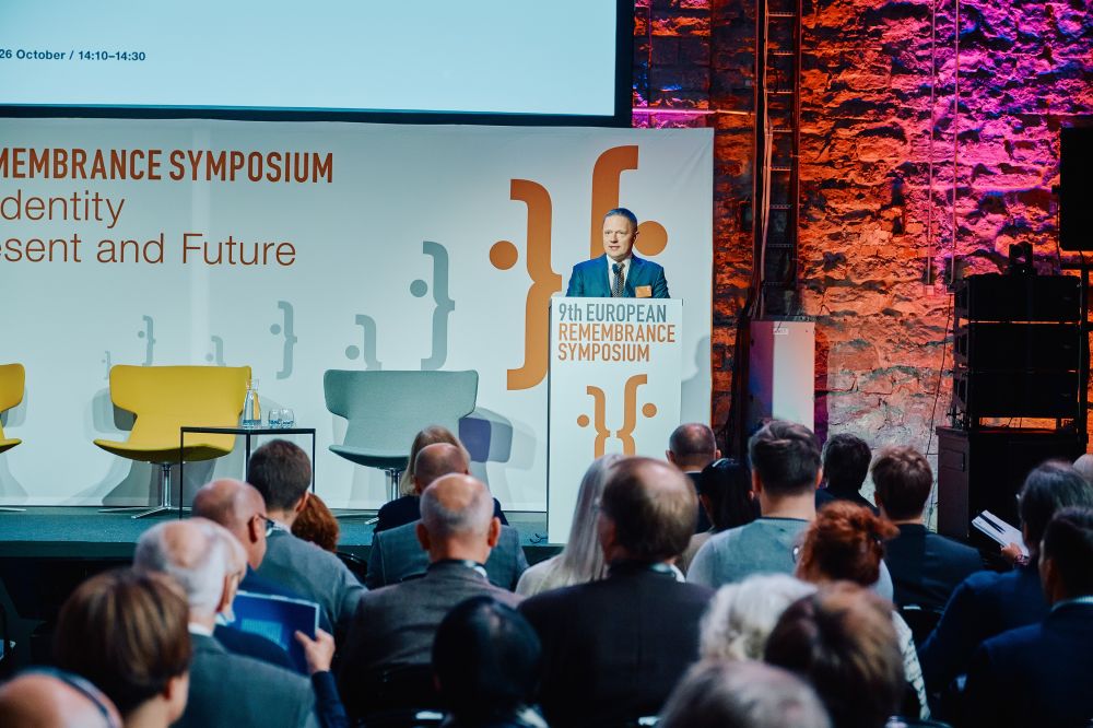 Interior, large postindustrial hall. Rafał Rogulski, ENRS Director, facing the camera speaks in front of a packed auditorium. He stands behind the lectern on a spacious stage, with the Ninth European Remembrance Symposium written on a board behind him.