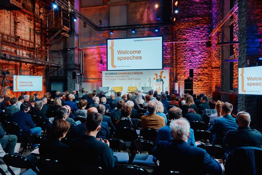 Photo taken from behind the audience. Large postindustrial hall. Rafał Rogulski, ENRS Director, facing the camera speaks in front of a packed auditorium. He stands behind the lectern on a spacious stage, with the Ninth European Remembrance Symposium written on a board behind him.