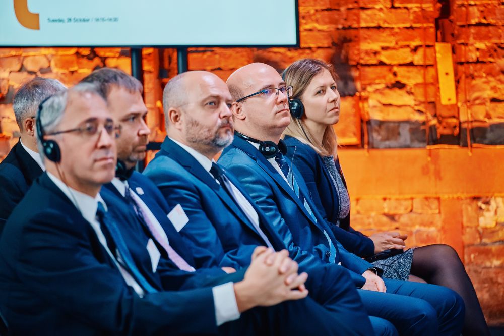 Interior, orange-lit postindustrial hall. A row of well-dressed attendees, four men and a woman, sits in an auditorium. They are listening attentively. Some of them are wearing simultaneous translation headphones.