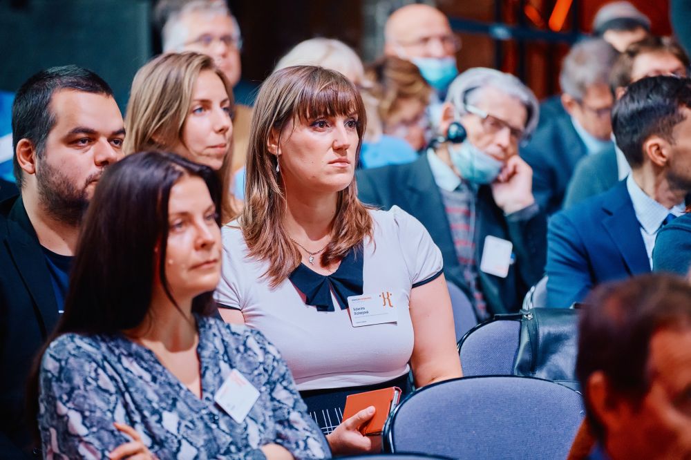 Interior, postindustrial hall. An auditorium full of well-dressed attendees listens attentively to the speaker. They are sitting in multiple rows of black and blue folding chairs.