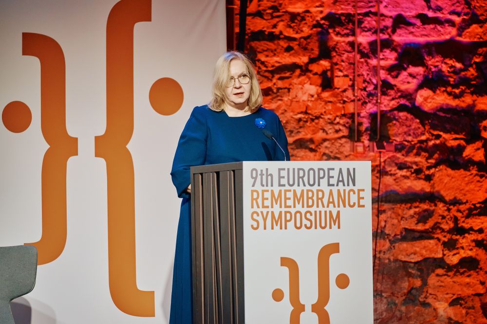 Interior, large postindustrial hall. Maris Lauri, Minister of Justice of Estonia, speaks in front of a packed auditorium. She is facing the camera, standing behind the lectern on a spacious stage, with the Ninth European Remembrance Symposium written on a board behind her.