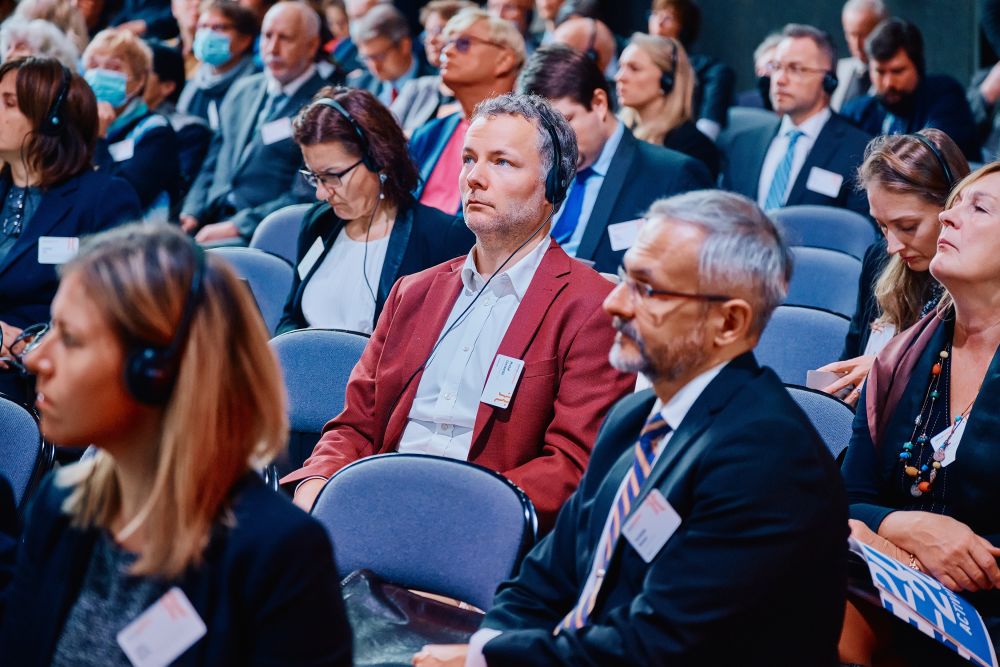 Interior, postindustrial hall. An auditorium full of well-dressed attendees listens attentively to the speaker. In the center of the photo, Dr MichaŁ Łuczewski seats in a dark-red jacket.