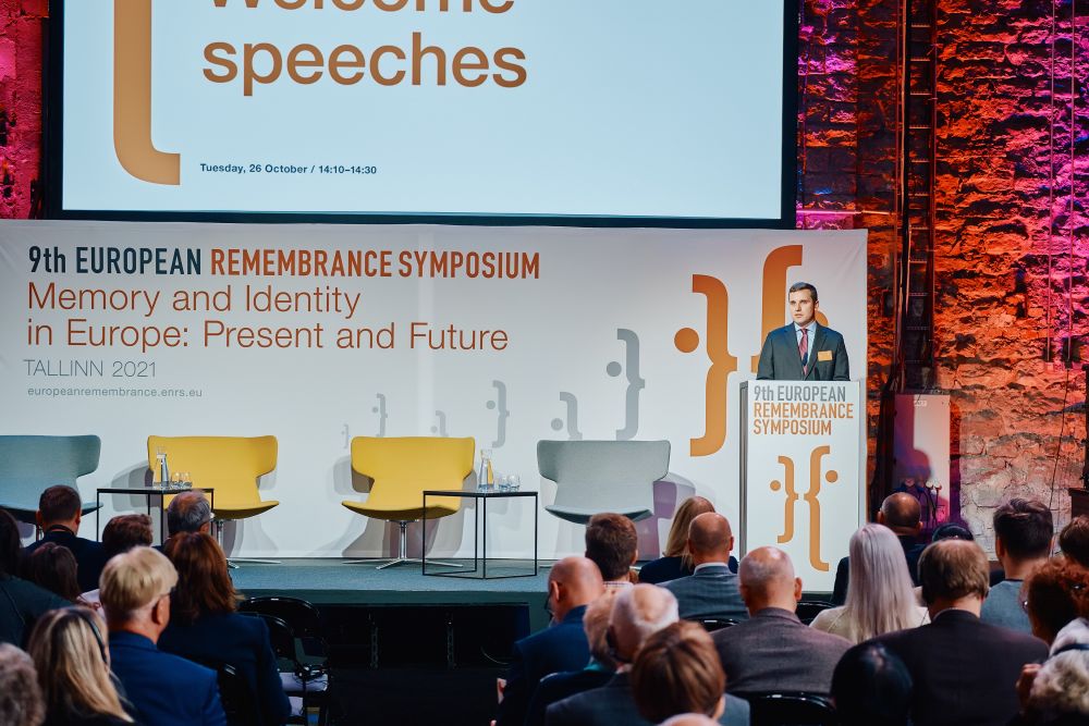 Interior, large postindustrial hall. Sergei Metlev, board member of the  Estonian Institute of Historical Memory speaks in front of a packed auditorium. He is facing the camera, standing behind the lectern on a spacious stage, with the Ninth European Remembrance Symposium written on a board behind him.