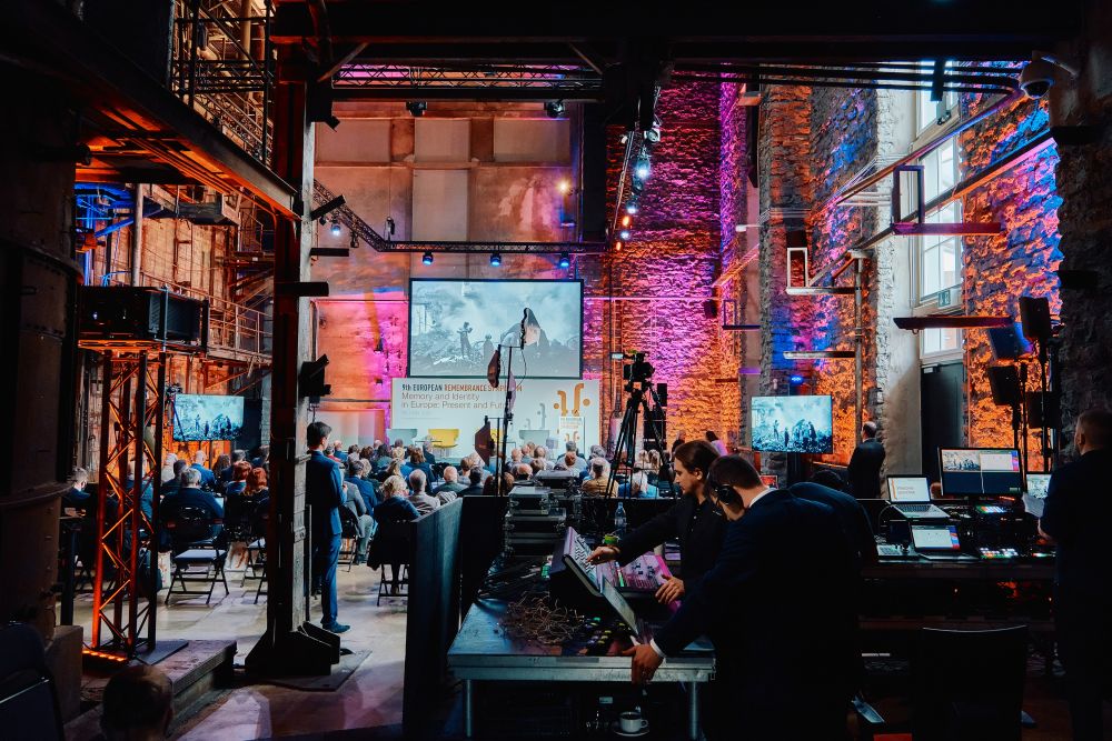 Interior, large postindustrial hall. In the foreground, a technical table with a stage engineering setup. Two stage engineers are busy with one of the control panels. In the background, the rows of chairs of the auditorium and a stage of the Ninth European Remembrance Symposium