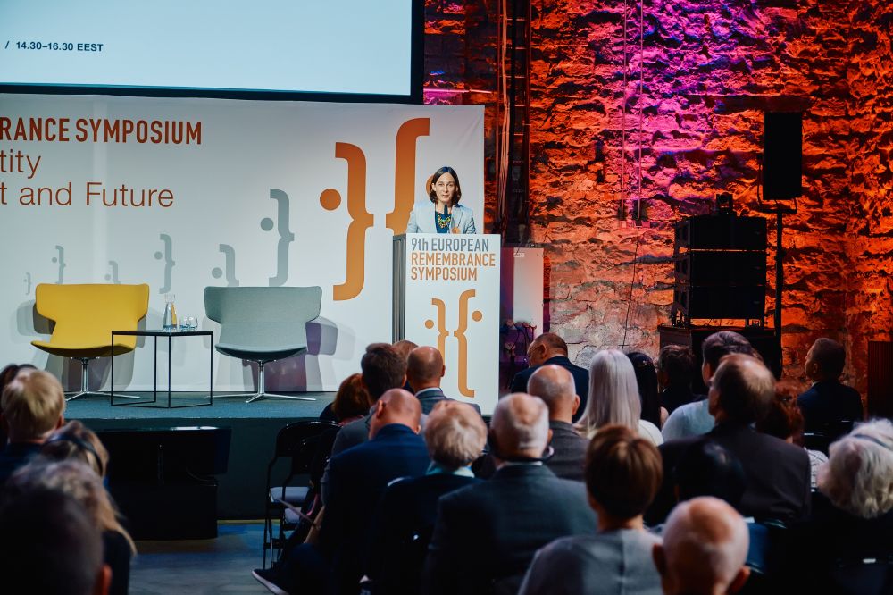 Interior, large postindustrial hall. A woman speaks in front of a packed auditorium. She is facing the camera, standing behind the lectern on a spacious stage, with the Ninth European Remembrance Symposium written on a board behind her.