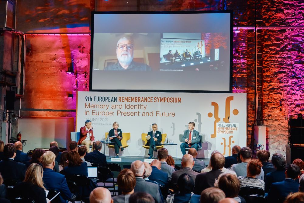 Dr Michał Łuczewski, Dr Monika Kareniauskaitė,  Prof. Viacheslav Morozov, and Toomas Hiio sit in the chairs placed on a stage. This panel discussion is joined online by Prof. Elazar Barkan, whose camera feed is projected onto a screen above the panelists. In front of the stage, a full auditorium.