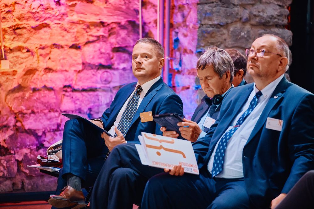 Interior, orange-lit postindustrial hall. A row of well-dressed attendees sits in an auditorium. They are listening attentively. Among them, Rafał Rogucki, ENRS Director, and Profesor Jan Rydel.