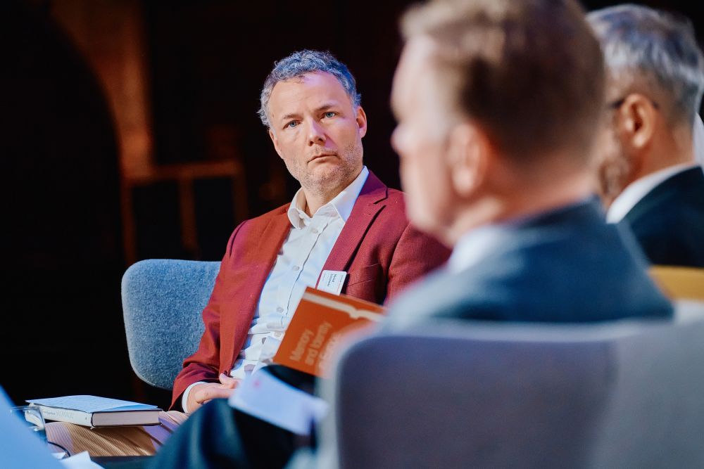 The main stage during a discussion panel. In the cameras focus, Dr Michał Łuczewski sits in a chair, listening with slight surprise to his co-panelist. Out of the focus, other panel participants.