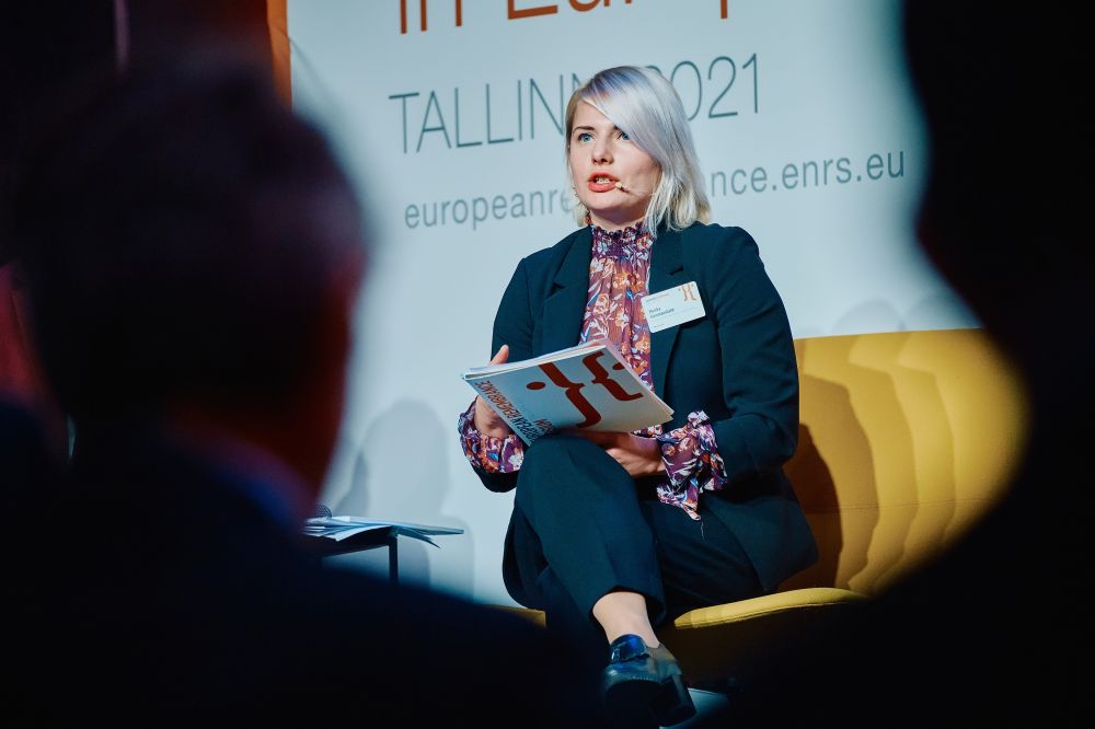 The main stage during a discussion panel. Dr Monika Kareniauskaitė sits in a yellow chair, speaking to a microphone addressing the audience. In front of her, out of focus, the silhouettes of the listeners.
