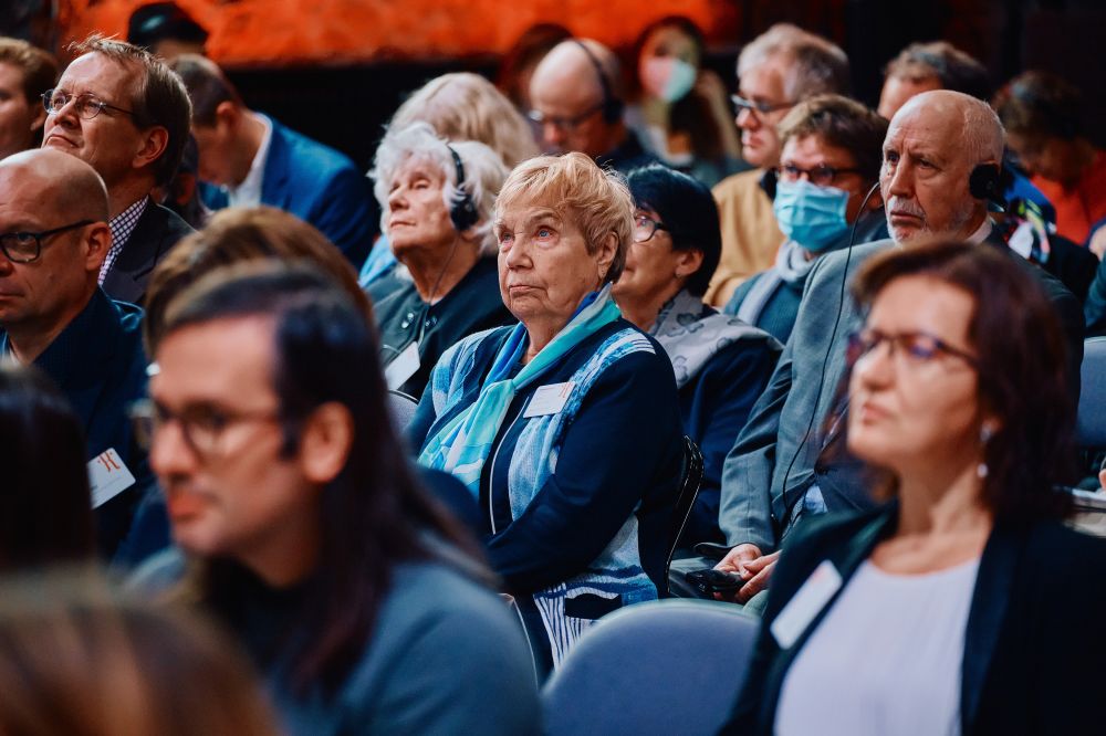 Interior, postindustrial hall. An auditorium full of well-dressed attendees listens attentively to the speaker. They are sitting in multiple rows of black and blue folding chairs.