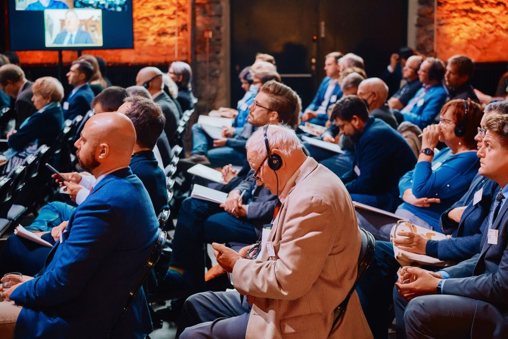 Interior, orange-lit postindustrial hall. Rows of well-dressed attendees sit in an auditorium, facing the stage on the left. They are listening attentively. Some of them are wearing simultaneous translation headphones.