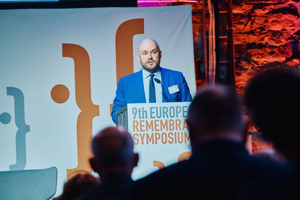 Interior, large postindustrial hall. Martin Andreller speaks in front of an auditorium. He is facing the camera, standing behind the lectern on a spacious stage, with the Ninth European Remembrance Symposium written on it.