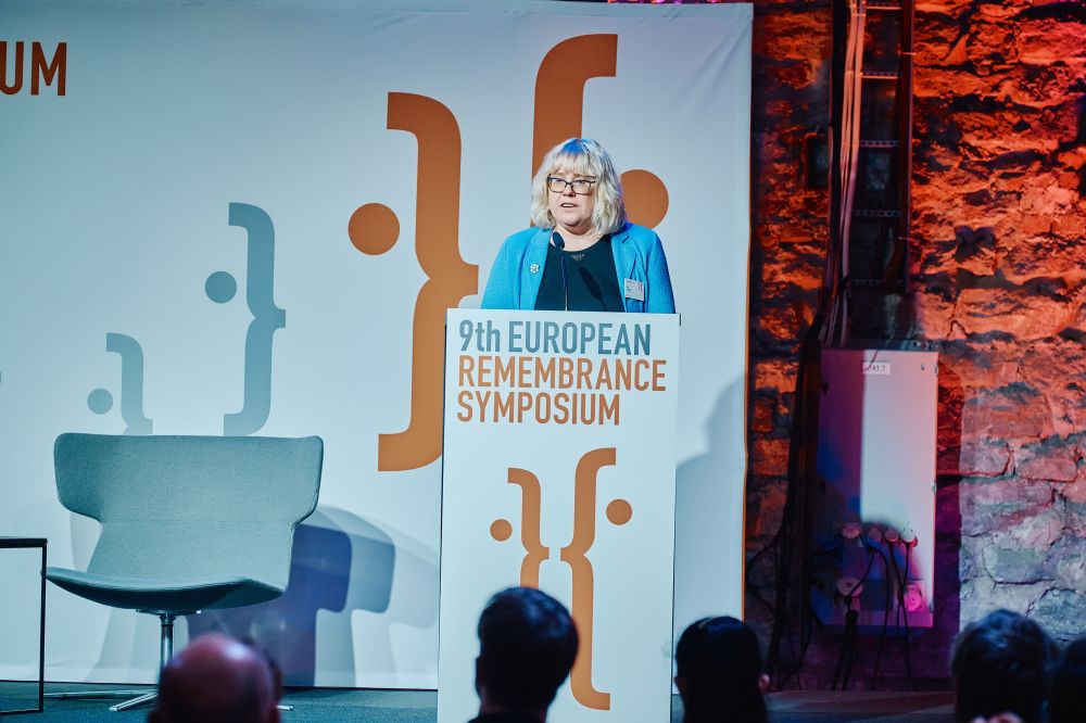 Interior, large postindustrial hall. Terje Anepaio speaks in front of an auditorium. She is facing the camera, standing behind the lectern on a spacious stage, with the Ninth European Remembrance Symposium written on it.