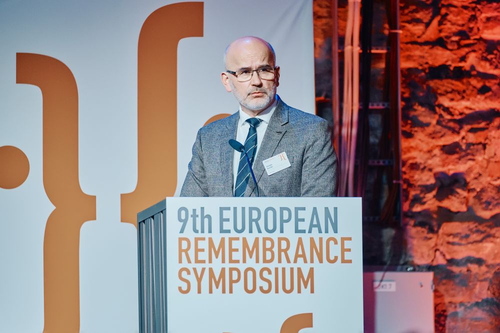 Interior, large postindustrial hall. Grzegorz Berendt speaks in front of an auditorium. He is facing the camera, standing behind the lectern on a spacious stage, with the Ninth European Remembrance Symposium written on it.