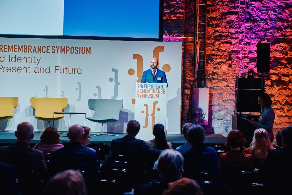 Interior, large postindustrial hall. Murray Bessette speaks in front of an auditorium. He is facing the camera, standing behind the lectern on a spacious stage, with the Ninth European Remembrance Symposium written on it.