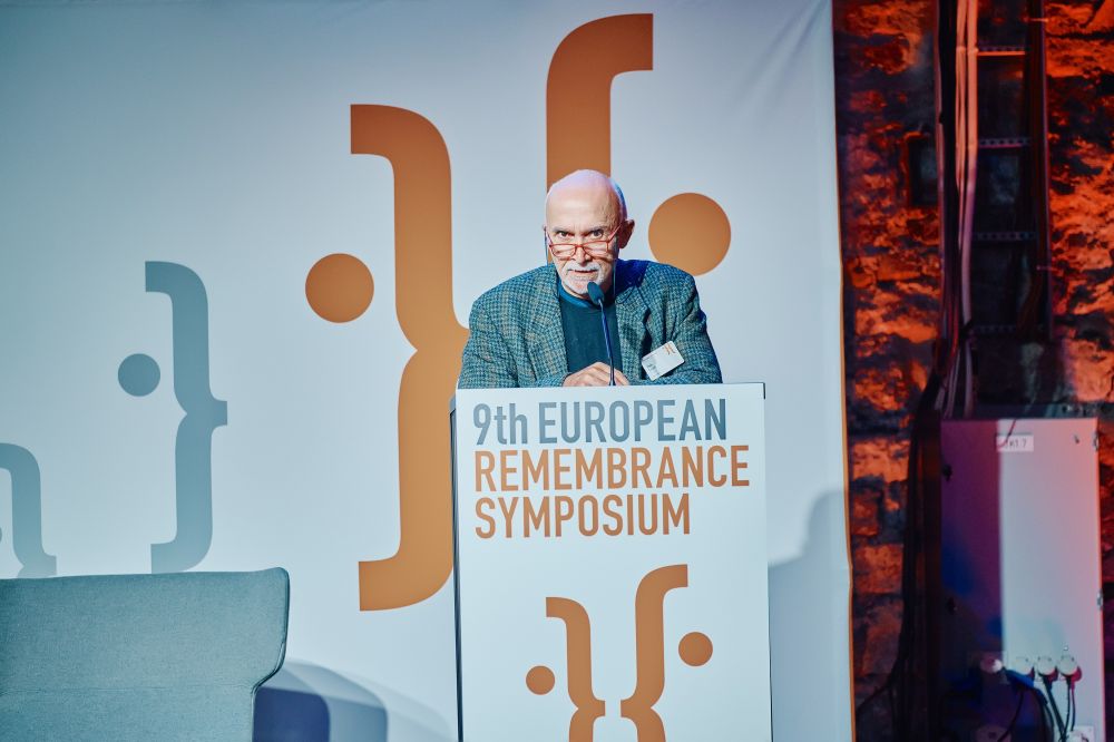 Interior, large postindustrial hall. Paolo Ceccoli stands behind the lectern on the lectern on a spacious stage, with the Ninth European Remembrance Symposium written on it.