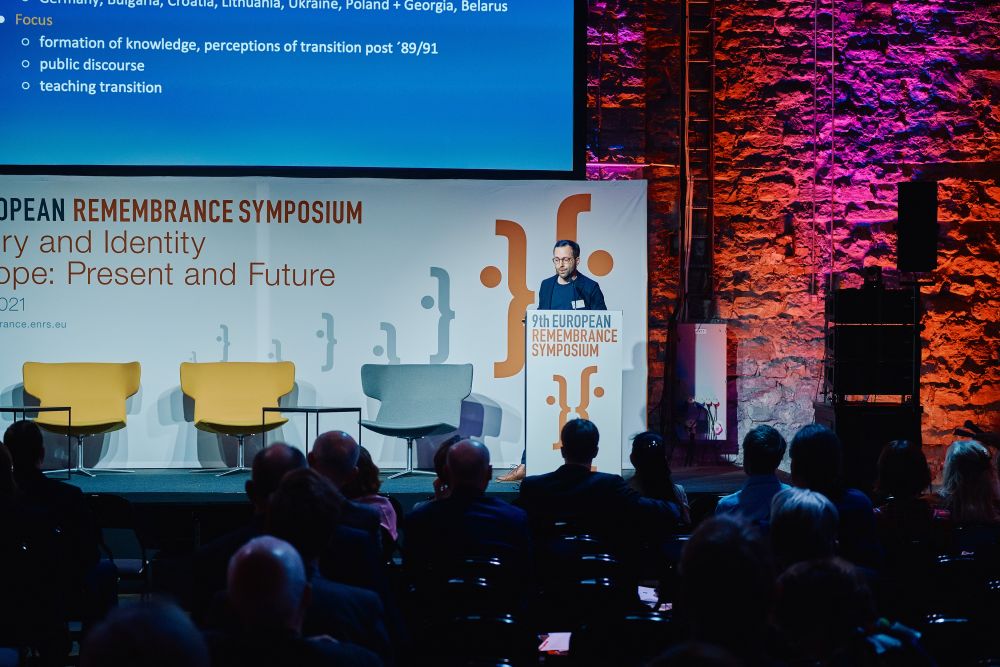 Interior, large postindustrial hall. Alexander Formozov speaks in front of an auditorium. He is facing the camera, standing behind the lectern on a spacious stage, with the Ninth European Remembrance Symposium written on it.