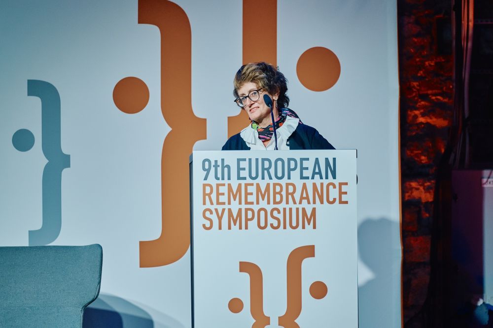 Interior, large postindustrial hall. Elżbieta Moczarska speaks in front of an auditorium. She is facing the camera, standing behind the lectern on a spacious stage, with the Ninth European Remembrance Symposium written on it.