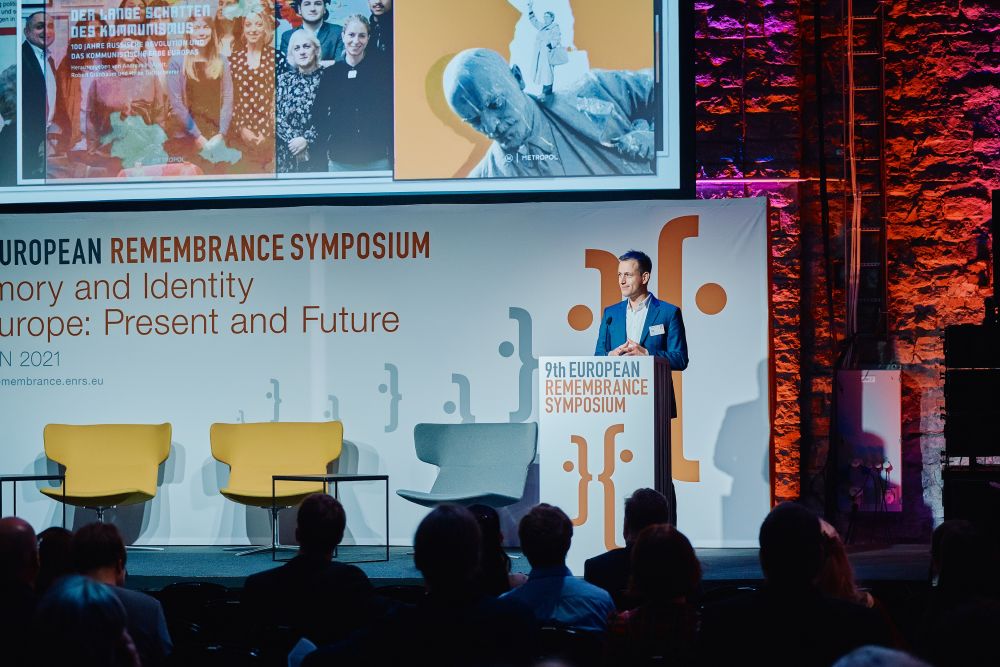 Interior, large postindustrial hall. Vincent Regente speaks in front of an auditorium. He is standing behind the lectern on a spacious stage, with the Ninth European Remembrance Symposium written on it. Above him, a multimedia presentation is displayed on a screen.