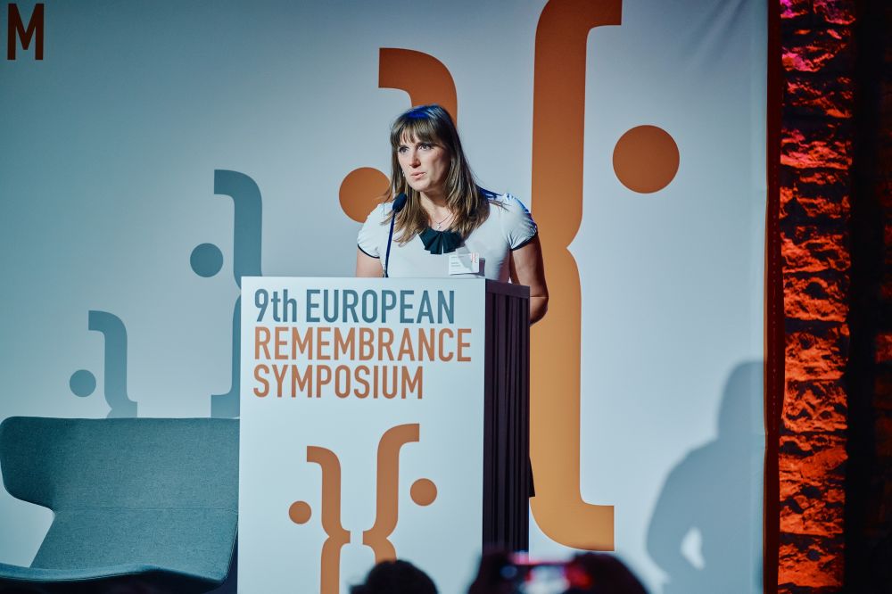 Interior, large postindustrial hall. Katarina Ristveyová speaks in front of an auditorium. She is facing the camera, standing behind the lectern on a spacious stage, with the Ninth European Remembrance Symposium written on it.