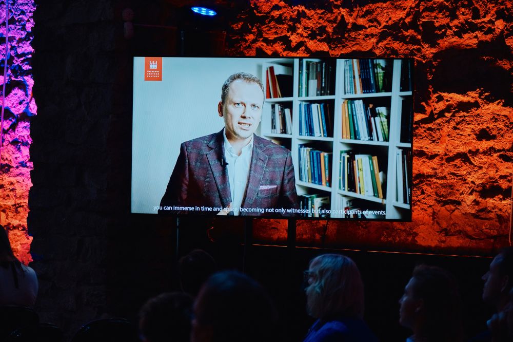 Interior, large postindustrial hall.  A large screen hangs from the wall next to the stage. Displayed on it, Rafał Wiśniewski stands in front of a bookshelf,  addressing the auditorium gathered in Tallinn.