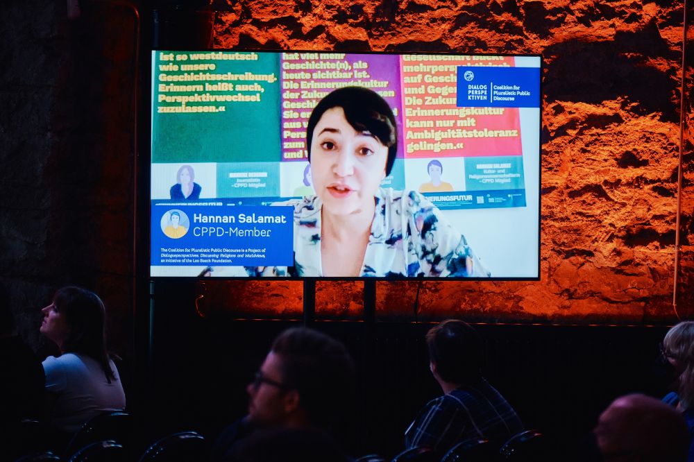 Interior, large postindustrial hall.  A large screen mounted to the orange-lit wall next to the stage. Displayed on it, Hannan Salamat, CPPD member, addresses the auditorium gathered in Tallinn.