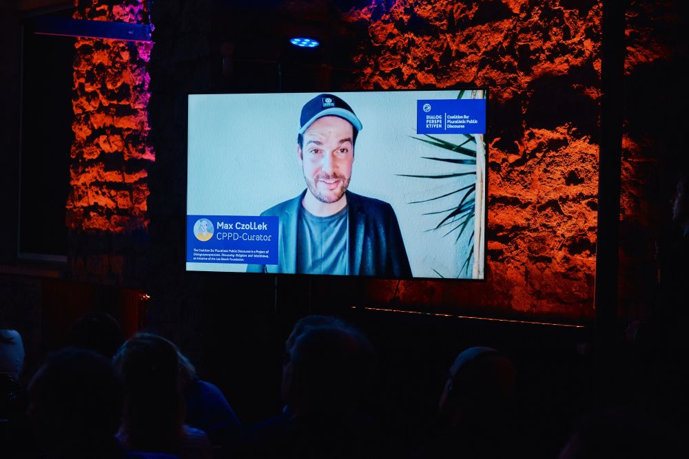 Interior, large postindustrial hall.  A large screen mounted to the orange-lit wall next to the stage. Displayed on it, Max Czollek, CCPD curator, addresses the auditorium gathered in Tallinn.