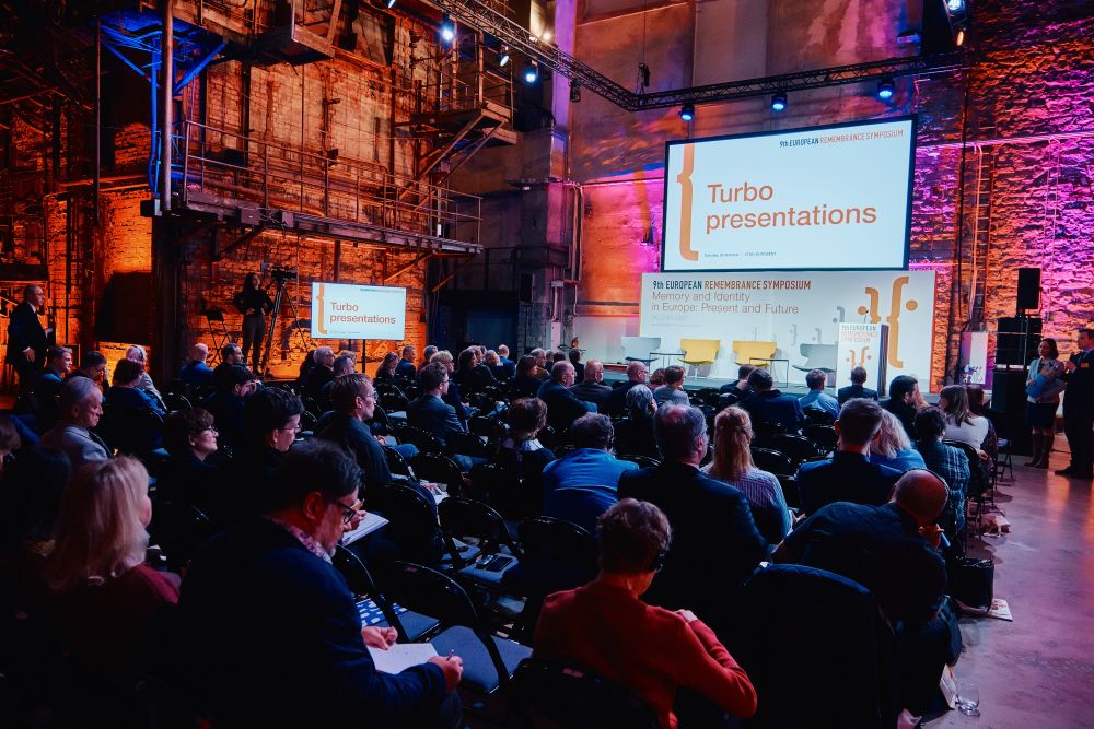 Interior. A spacious postindustrial hall is illuminated with orange lights. Multiple rows of occupied auditorium chairs. In the background, still empty stage. Above the lectern, a  title of the incoming session, named Tubo presentations, is displayed on a large screen.