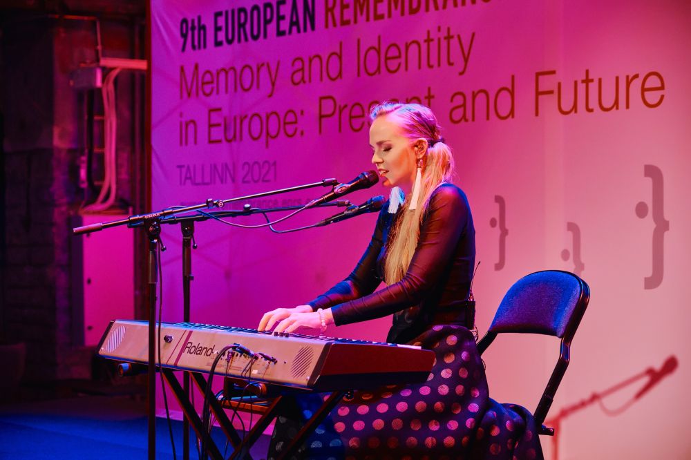 Interior, a pink-lit scene in the large postindustrial hall. Kadi Voorand sits in front of an electric piano. She plays the instrument, singing into a microphone standing in front of her. In the background, a banner with the Ninth European Remembrance Symposium written on it.