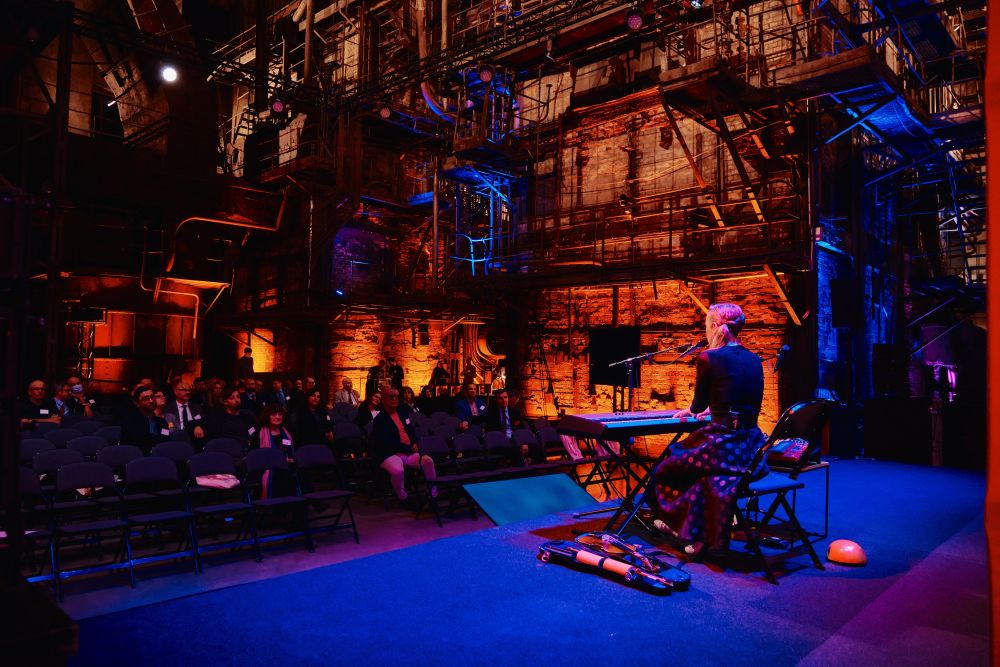 Interior, an orange-lit large postindustrial hall. Kadi Voorand sits in front of an electric piano, facing back the camera. She plays the instrument, singing into a microphone standing in front of her. In front of her, rows of sited in a dark audience.