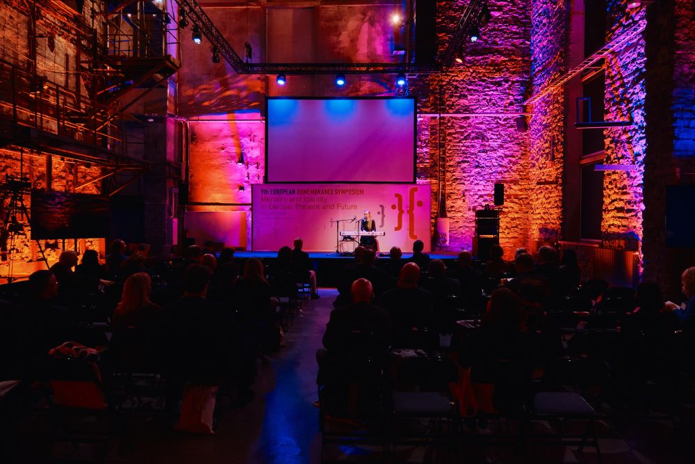 Interior, a pink-lit scene in the large postindustrial hall. Kadi Voorand sits in front of an electric piano. She plays the instrument, singing into a microphone standing in front of her. In front of her, a large audience.