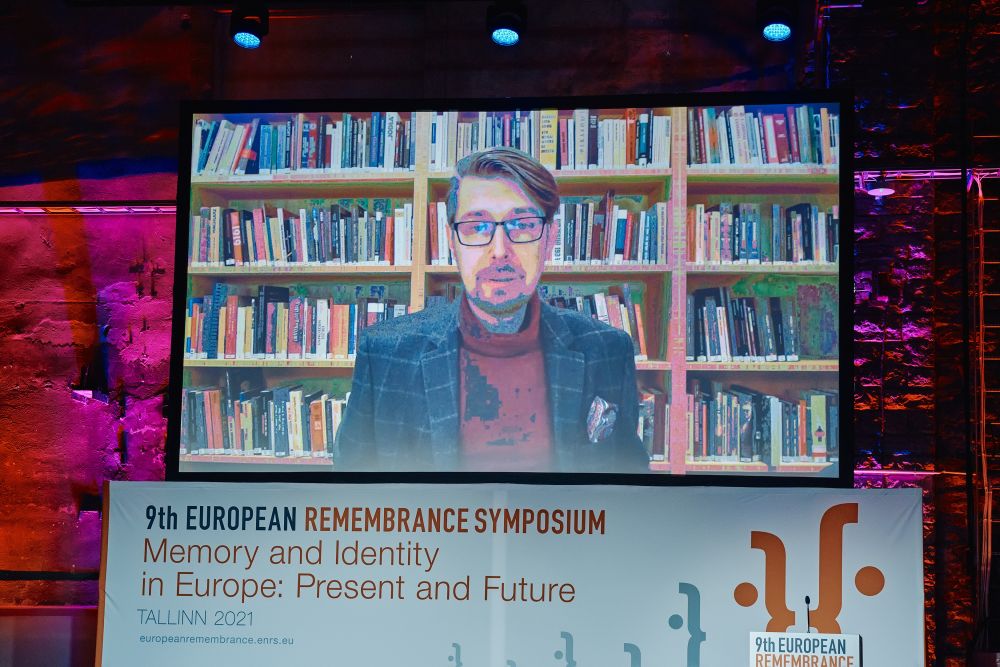 Interior, large postindustrial hall. a large pull-down projector screen hangs above the empty stage. Displayed on it, Łukasz Jasiński adresses the auditorium gathered in Tallinn.