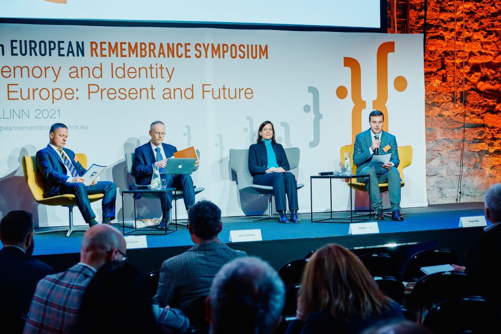 Rafał Rogulski, Dr Łukasz Kamiński,  Constanze Itzel, and Sergei Metlev sit in the chairs placed on a stage. Behind them, a banner with the Ninth European Remembrance Symposium written on it. In front of them,  rows of the auditorium.